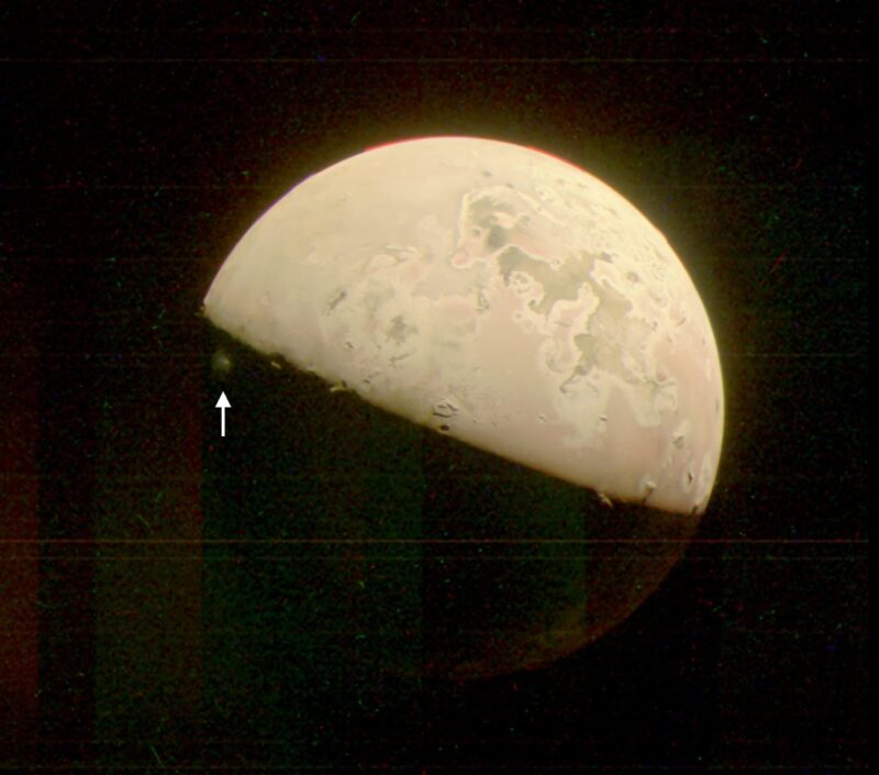 ジュノー探査機がジュノーカムでとらえた木星の衛星イオ。2023年10月15日にイオに接近した際に撮影されました。昼夜境界の下側（白い矢印）に、プロメテウス火山の噴煙が映っています。